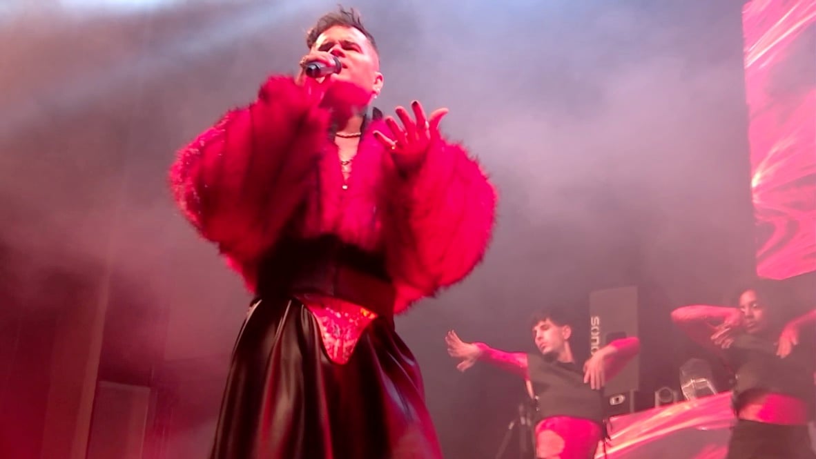 Lui da Motta con abrigo de pelo rojo, brazos en alto, bajo luz rosa.